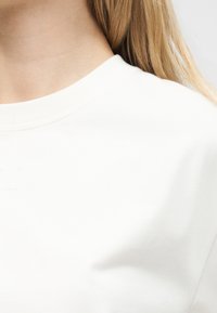 Κοντινό πλάνο του λαιμού και του ώμου ενός ατόμου με μακριά ξανθά μαλλιά, φορώντας ένα απλό λευκό πουκάμισο με στρογγυλή λαιμόκοψη.