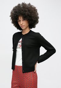 Sort struktureret cardigan med rund hals og knaplukning, båret over en hvid skjorte med grafik, kombineret med mønstrede røde bukser.