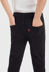 Pantalones de mezclilla negros con dos bolsillos traseros, con detalles de costura y una etiqueta roja con logo. La tela tiene una textura suave y un corte clásico.