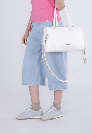 Persona con jeans azzurri a gamba larga e top rosa, che tiene una borsa bianca con tracolla, indossa scarpe da ginnastica grigio chiaro e calzini beige.