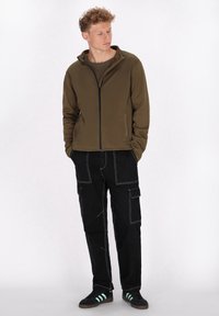 Olijfgroene zip-up jack met hoge kraag, zwarte cargo broek met witte stiksels en zwarte sneakers met groene strepen.