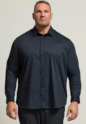 Hombre musculoso con cabello corto y barba que lleva una camisa azul marino oscura de manga larga con botones, de pie frente a un fondo claro y liso.