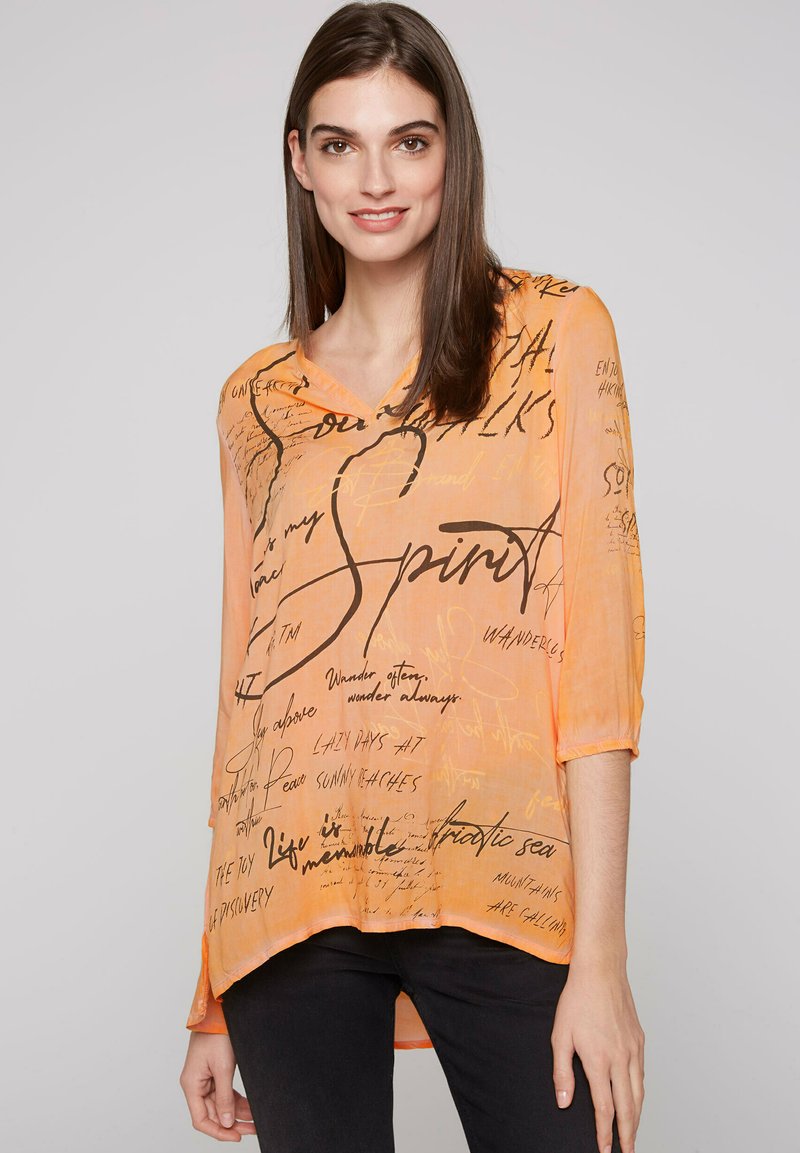 Helles orangefarbendes, durchscheinendes Blusenshirt mit schwarzem, handschriftlichem Text und Mustern. Mit V-Ausschnitt, 3/4-Ärmeln und einem asymmetrischen Saumdesign.