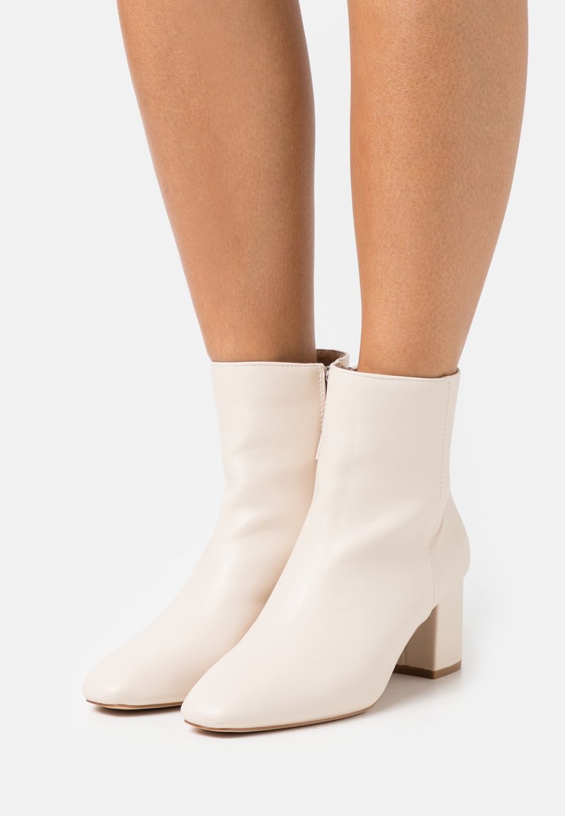 Bottines en cuir blanc cassé montant jusqu'à la cheville avec un talon carré, portées sur des jambes nues sur un fond uni.