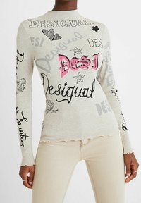 Top à manches longues en jersey léger gris clair avec des imprimés graphiques multicolores, incluant "DESI" en rose, des cœurs et des fleurs ; détail de l'ourlet ondulé.