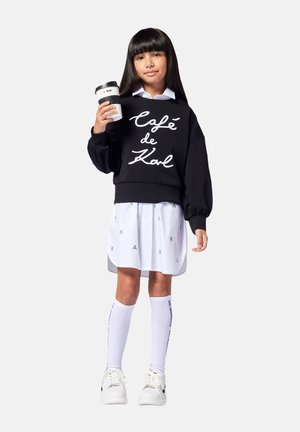 Felpa nera oversize con testo bianco "Café de Kal", gonna bianca con motivo grigio "KARL", calze bianche fino al ginocchio e sneakers bianche.