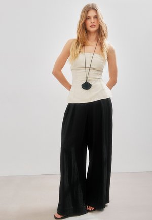 Žena s dlhými blond vlasmi, ktorá má na sebe telový top bez ramienok, široké čierne nohavice, čierny náhrdelník z mušlí a čierne sandále, stojí s rukami za chrbtom.