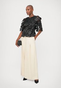 Model iført en sort blomsterdesignet gennemsigtig top, kremfarvede brede plisserede bukser, sorte spidse hæle, og holder en lille sort taske.