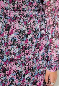 Robe en tissu noir avec un motif floral en rose, bleu et blanc. Présente une taille smockée et de longues manches bouffantes avec des poignets en élastique.