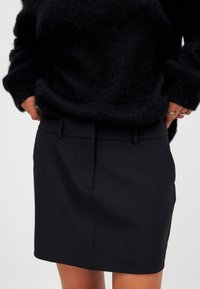 Gonna nera con un design sartoriale, caratterizzata da una vita piatta, tasche laterali e una texture di stoffa liscia. Adatta per diverse occasioni.