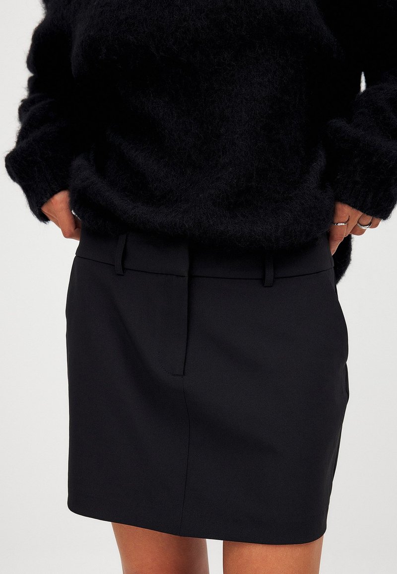 Gonna nera con un design sartoriale, caratterizzata da una vita piatta, tasche laterali e una texture di stoffa liscia. Adatta per diverse occasioni.