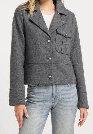 Žena v šedém vlněném saku na knoflíky s klopovou kapsou a vybledlých modrých džínách na bílém pozadí.