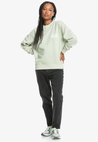 Mintgrønn sweatshirt med rund hals, strukturert bomullsmateriale og et emblem på venstre bryst. Kledd sammen med svarte jeans og sneakers.