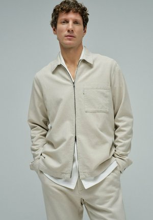 Hombre con cabello rizado que lleva una chaqueta beige con cremallera sobre una camisa blanca y pantalones beige a juego, de pie con las manos en los bolsillos.
