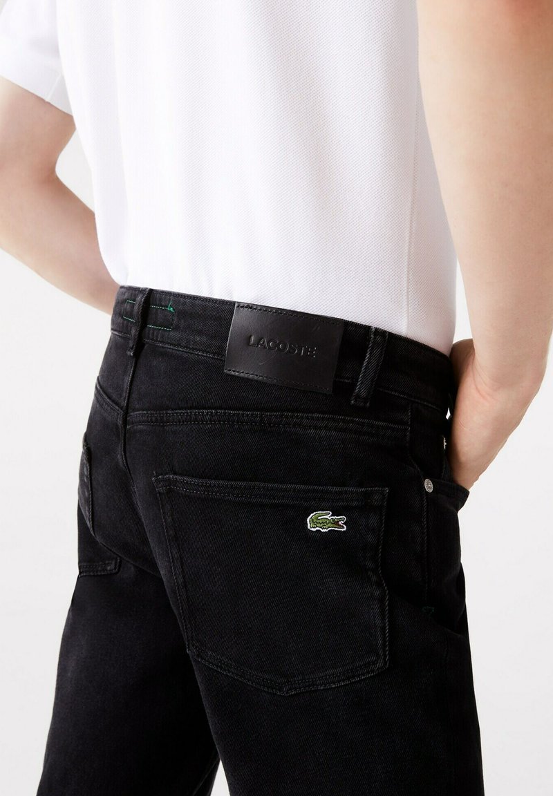 Schwarze Denim-Jeans mit einem Leder-Lacoste-Logo-Patch am Bund und einem grünen, gestickten Krokodil-Logo auf der Gesäßtasche.