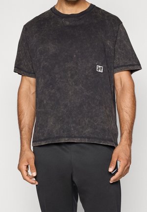 Mann trägt ein ausgewaschenes schwarzes T-Shirt mit kurzen Ärmeln und einem kleinen quadratischen Logo auf der linken Brust, kombiniert mit schwarzen Hosen.