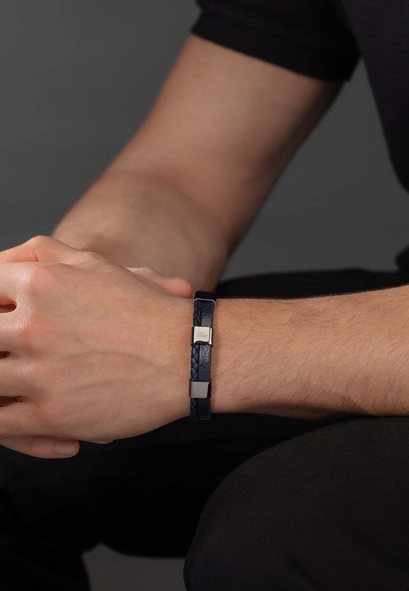 Marineblauwe gevlochten leren armband met metallic vierkante accenten, met een gestructureerde afwerking en een slank design, gedragen om een pols.