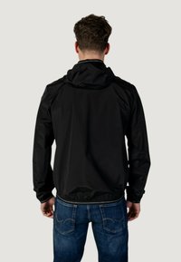 Giacca nera in nylon con cappuccio, polsini elastici e orlo. Indossata sopra jeans azzurri in denim, che mettono in evidenza una texture liscia e un design semplice.