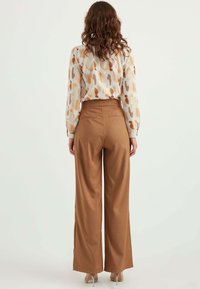 Leichte, gemusterte Bluse in Creme mit orangefarbenen und braunen Pinselstrich-Designs; hochtaillierte, weit geschnittene braune Hose mit Gesäßtaschen.