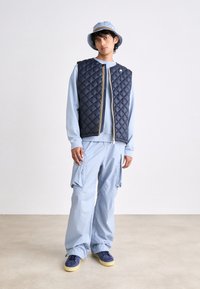 Navy quilted vest with yellow zipper, light blue sweatshirt, light blue cargo pants, and blue shoes with yellow soles. Bucket hat completes the look.
