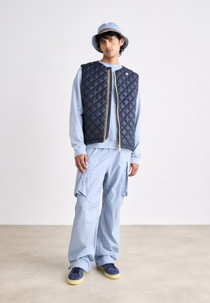 Chaleco acolchado navy con cremallera amarilla, sudadera azul clara, pantalones cargo azul clara, y zapatillas azules con suelas amarillas. Un sombrero tipo bucket completa el look.