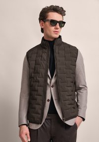 Dunkelolivfarbene Steppweste mit einem abgesteppten Muster, getragen über einem hellgrauen Blazer. Das Modell trägt schwarze Sonnenbrillen. Glatte, matte Textur.