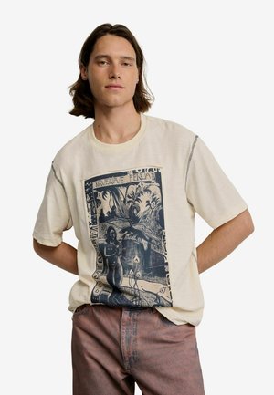 Junger Mann mit schulterlangem Haar, der ein beiges T-Shirt mit Grafik und ausgewaschene rosafarbene Hose trägt, steht mit den Händen hinter dem Rücken.