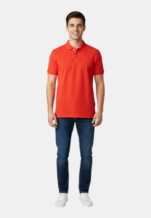 Giovane uomo in piedi con una polo arancione brillante, jeans blu e scarpe da ginnastica bianche su uno sfondo chiaro uniforme.