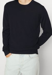 Granatowy sweter dziany z okrągłym dekoltem i długimi rękawami, z prążkowaniem na mankietach i dole, w zestawieniu z jasnoniebieskimi dżinsami.