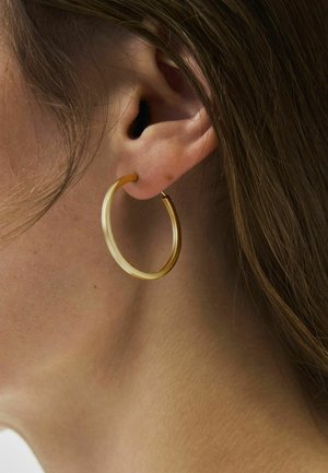 Prim-plan cu urechea stângă a unei persoane cu păr șaten deschis, purtând un cercel rotund, translucid, de culoare chihlimbar.