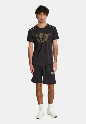 Uomo che indossa una maglietta sportiva nera e pantaloncini con logo "LEONE", calzini bianchi e scarpe da ginnastica bianche, in piedi su uno sfondo semplice.