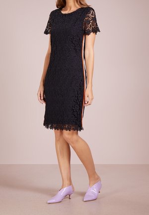 Vrouw gekleed in een zwarte kanten jurk tot op de knie met korte mouwen en zijstrepen, gecombineerd met lavendelkleurige pumps met een lage hak en puntige neus.