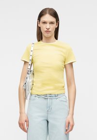 Mujer con cabello castaño liso que lleva una camiseta amarilla de manga corta y jeans azul claro, con un bolso plateado colgado del hombro.