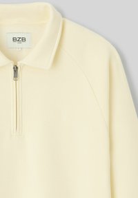 Pull cr�ème à manches longues avec fermeture éclair quart et col, présentant un petit logo discret sur la poitrine et une étiquette de marque BZB à l'intérieur.