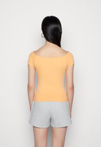 Πορτοκαλί διάφανο τοπ χωρίς ώμους, φτιαγμένο από μαλακό υλικό, εφαρμοστή σχεδίαση. Συνδυάζεται με ανοιχτό γκρι σορτσάκι. Το φόντο είναι απλό λευκό.