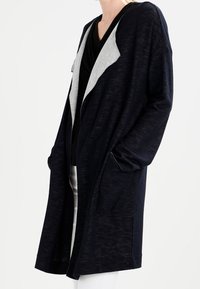 Cardigan lungo di un blu navy scuro con cappuccio e colletto grigio chiaro. Presenta due tasche frontali e una vestibilità relax, oversize. Tessuto morbido e strutturato.
