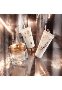 Three skincare products on a reflective surface: a glass jar with a gold lid, and two tubes in white with gold accents, featuring textured labels.