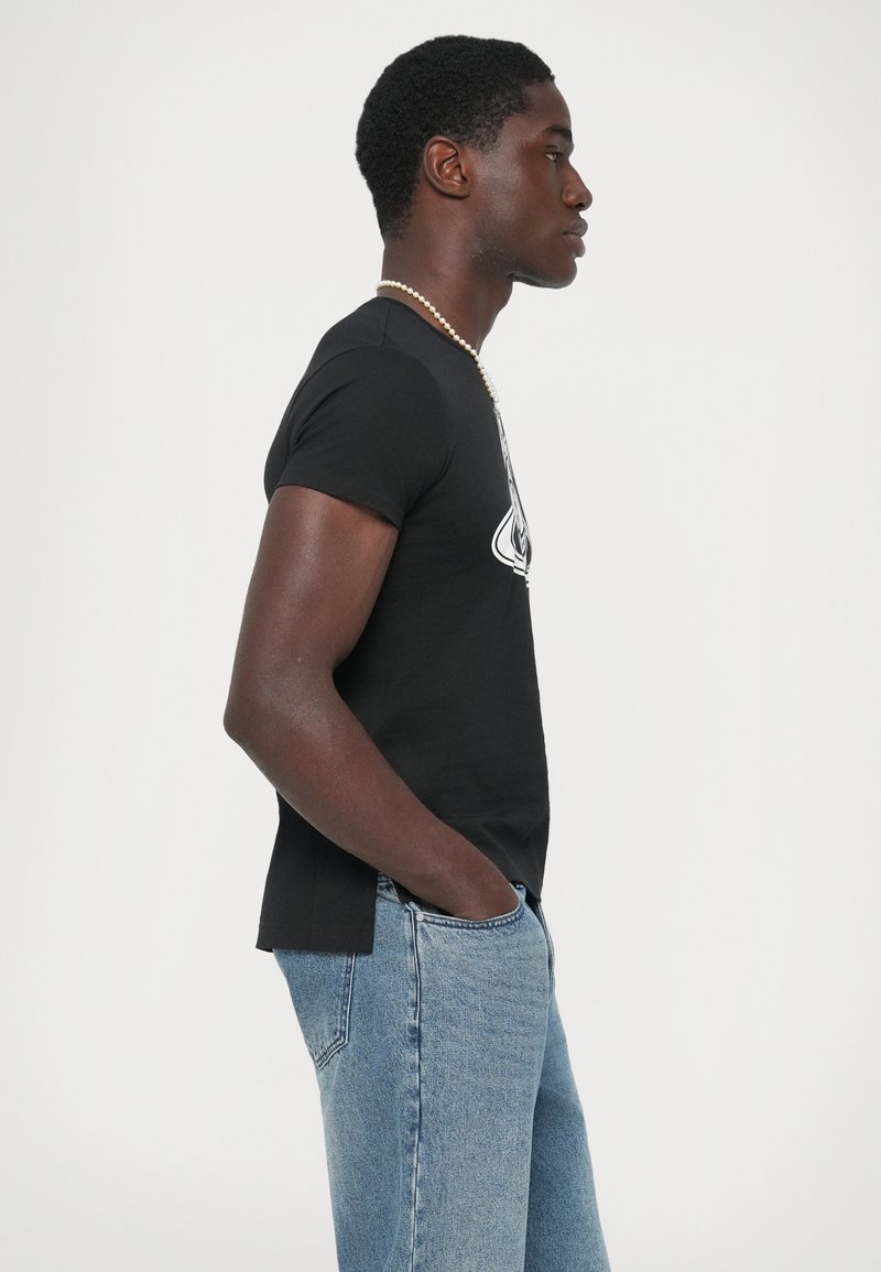 Uomo in t-shirt grafica nera, jeans azzurri chiari con le mani in tasca, che indossa una collana di perle, in profilo su uno sfondo bianco.