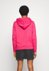 Sudadera con capucha de color rosa brillante y textura suave, con puños y dobladillo acanalados, vista desde la parte posterior, combinada con una falda de mezclilla negra.
