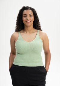 Hellgrünes geripptes Tanktop mit V-Ausschnitt und dünnen Trägern. Kombiniert mit schwarzen Hosen wirkt der Stoff weich und hat einen figurbetonten Schnitt.