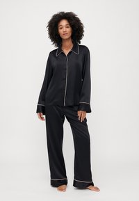Black satin pajama set with long sleeves and pants, featuring contrasting cream piping along the collar and cuffs. Smooth texture, relaxed fit.
