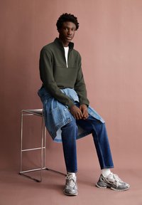 Felpa zipata verde scuro, t-shirt bianca, giubbotto di jeans annodato in vita, jeans blu e scarpe da ginnastica argento con calze bianche.