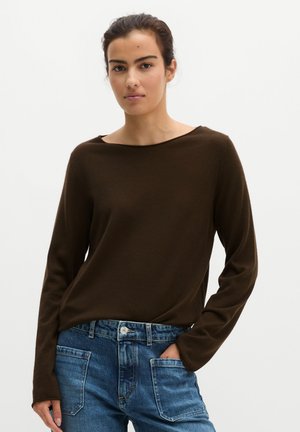Junge Frau mit dunklen zurückgebundenen Haaren trägt ein dunkelbraunes langärmliges Oberteil und blaue Jeans mit vorderen aufgesetzten Taschen, eine Hand in der Tasche.