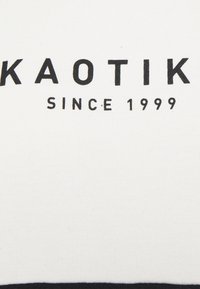 Bílá bavlněná látka s černým nápisem "KAOTIK" nad "SINCE 1999". Jednoduchý, výrazný design bez dalších vzorů nebo akcentů.