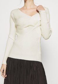 Sweter w kolorze kremowym z prążkowanym wzorem, w dekolcie w kształcie litery V i detalem w formie skrętu, noszony z plisowaną, ciemnobrązową spódnicą, z długimi rękawami i dopasowanym krojem.