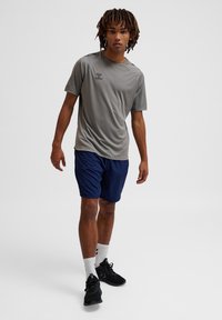 Jonge man loopt vooruit, gekleed in een grijs sportshirt, marineblauwe korte broek, witte sokken en zwarte sportschoenen op een witte achtergrond.