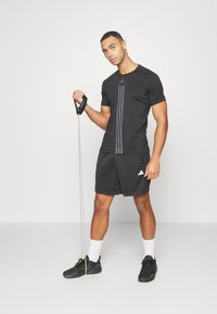 Sort sports T-shirt med tre lodrette grå striber, med sorte shorts og sorte sneakers, der holder en modstandsbånd.