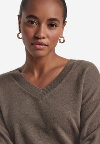 Pullover mit V-Ausschnitt und langen Ärmeln in einem gedämpften Braunton. Weiche Textur mit geripptem Ausschnitt. Model trägt goldene Creolen. Neutraler Hintergrund.