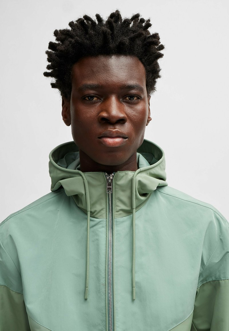 Hellgrüne Kapuzenjacke mit Reißverschluss, Kordelzugakzenten und unterschiedlichen Texturen. Das Gesicht des Models ist sichtbar, mit kurzem, lockigem Haar.