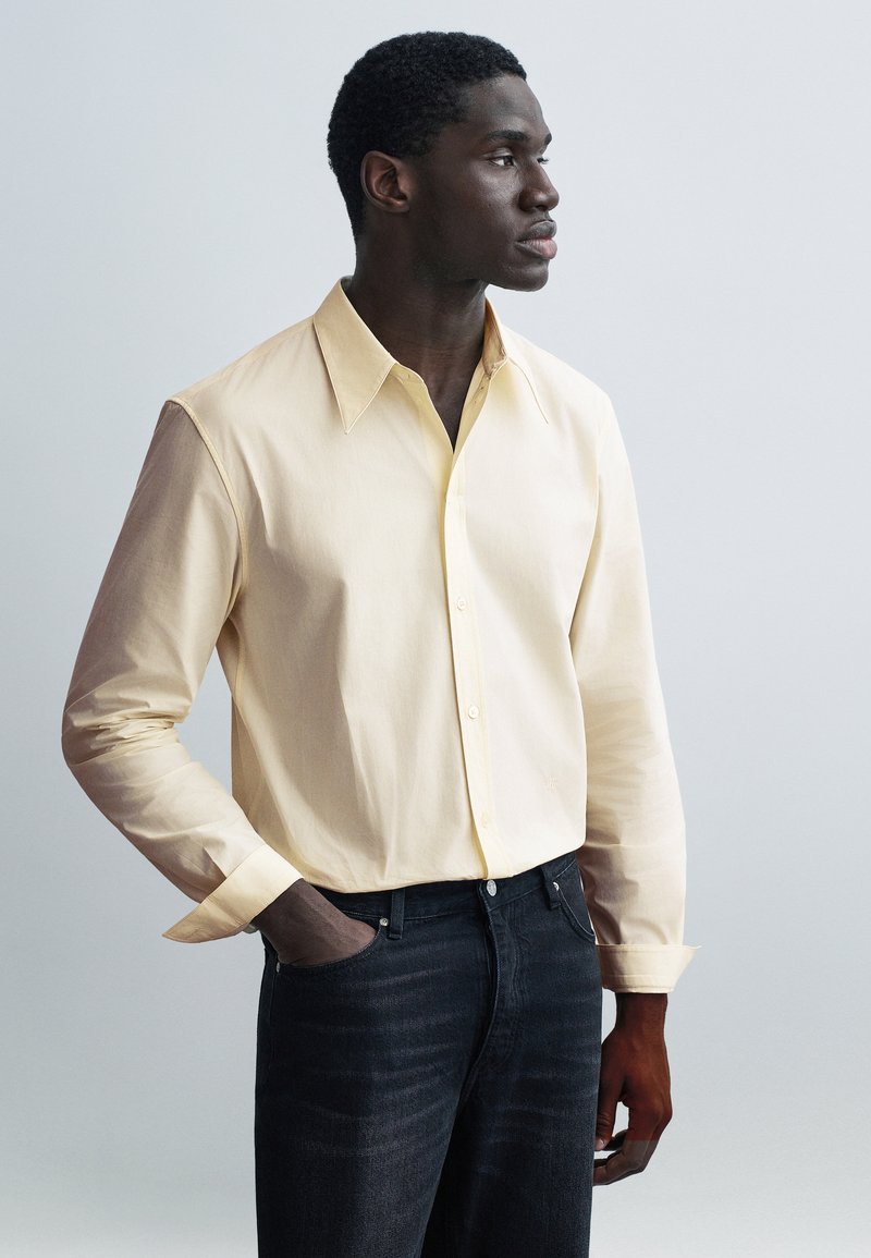 Jasnożółta koszula z długim rękawem, kołnierzem rozłożystym, zapinana na guziki z mankietami; w połączeniu z ciemnoniebieskimi dżinsami z denimu.